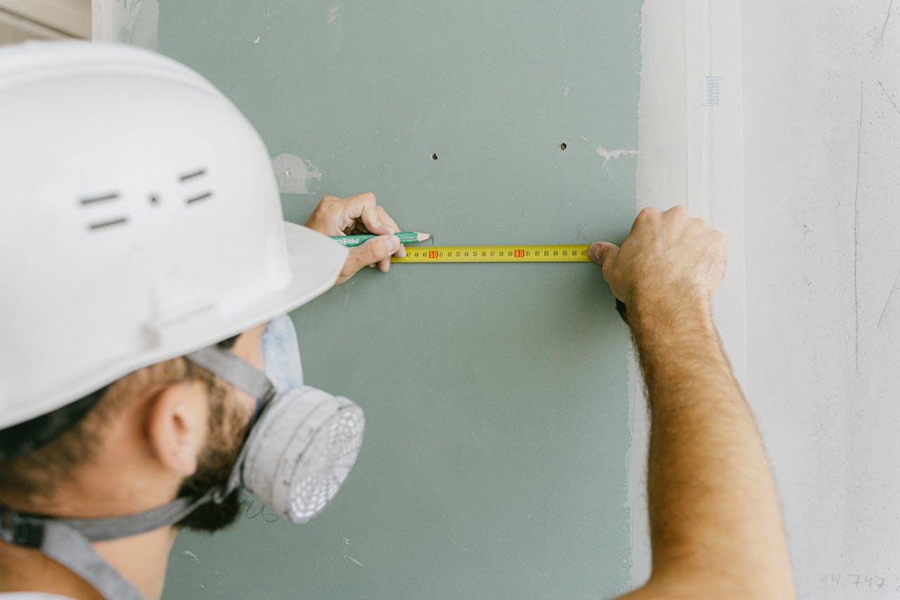 A construction worker precisely measuring drywall, wearing a helmet and mask for safety.